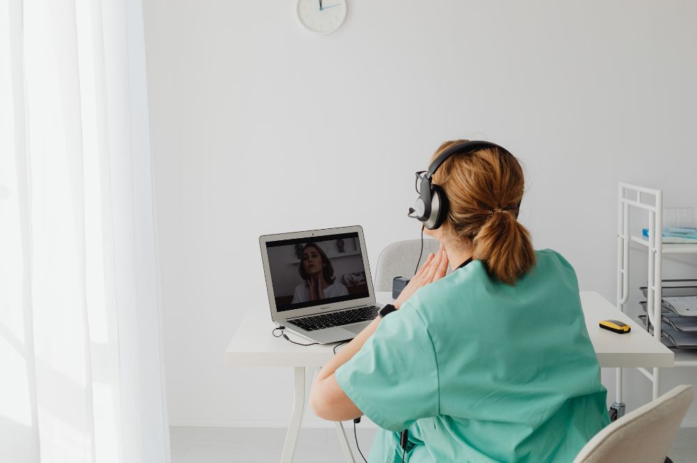 A nurse wearing a headset performs a investigator site follow-up with a patient over video chat.