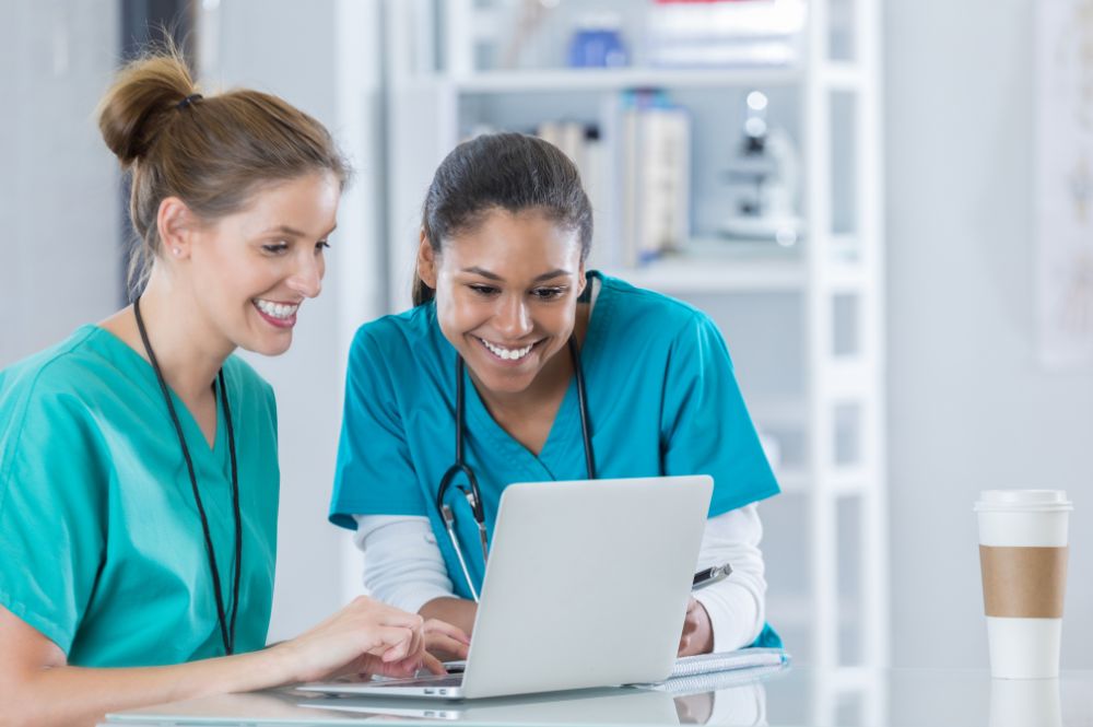 Two female nurses collaborate on a laptop during the secondary screening process.