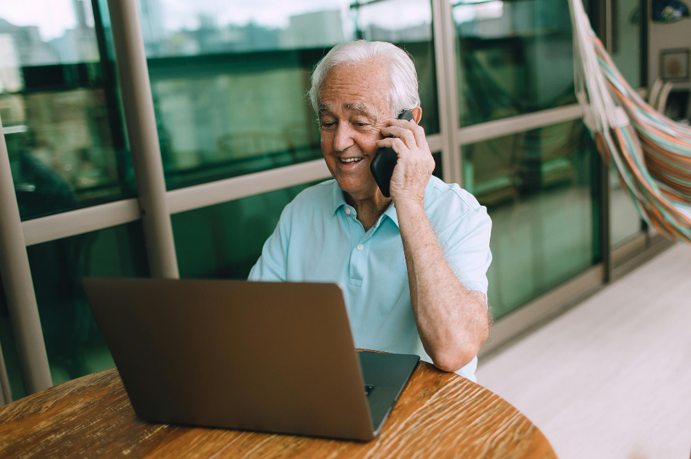 An older man at a table with a laptop, engaged in a phone conversation about patient engagement technology.